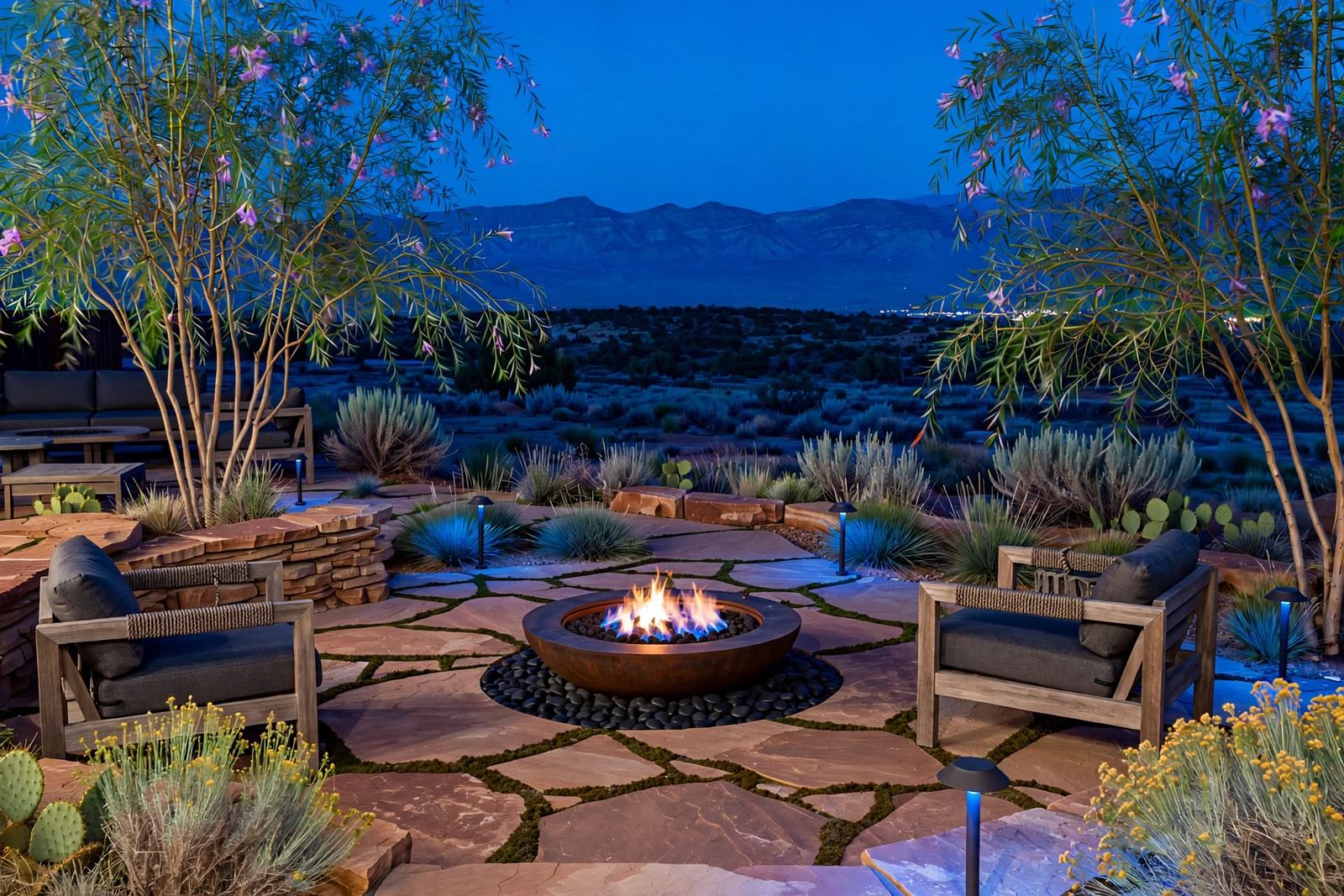 Flagstone patio with a lit fire pit, lounge chairs and softly uplit desert willows at blue hour